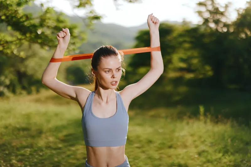 Young woman exercising with resistance band in nature, wearing sports apparel, demonstrating focus and determination, suitable for fitness and healthy lifestyle concepts