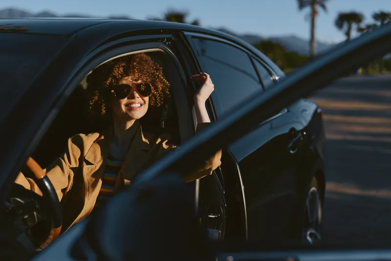 Casual woman inside a car, smiling in warm sunlight, a relaxed lifestyle moment with a retro mood, outdoors vignette, sunlit driving scene, natural tones and authentic joy