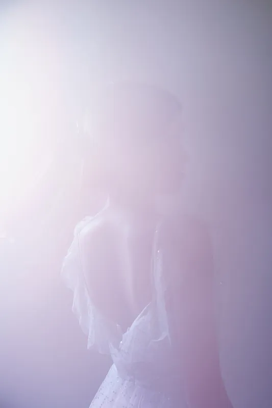 Elegant woman in a delicate white dress poses softly in diffused light. The image highlights beauty, fashion, and feminine grace with a dreamy, ethereal atmosphere.
