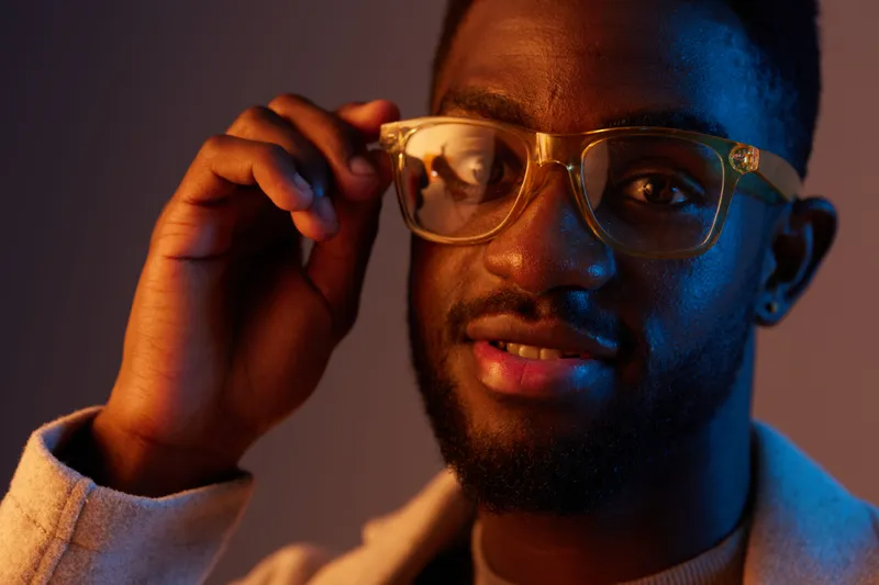 A stylish young African American man adjusting his glasses with a confident smile, illuminated by a warm blue orange background, showcasing modern fashion and self confidence