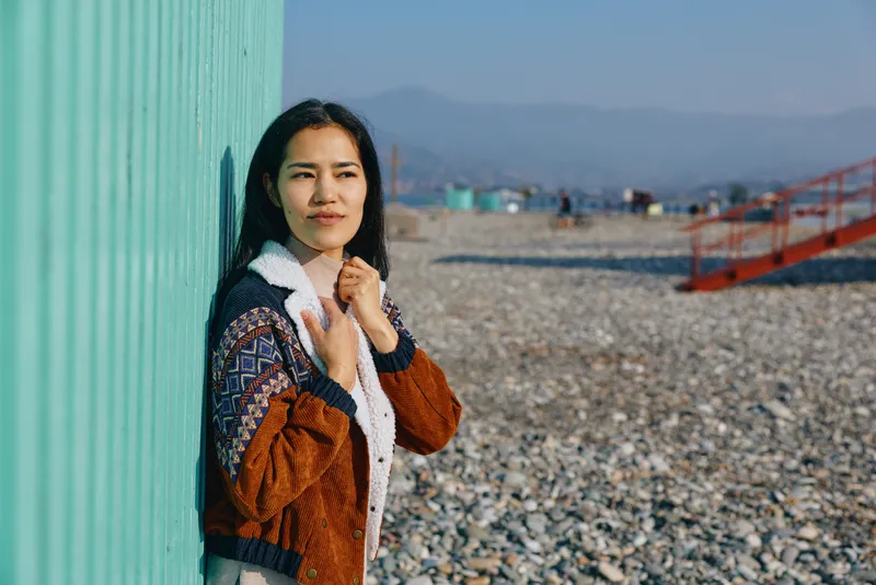 Woman in a patterned brown jacket leans against a turquoise wall on a pebbled beach, distant mountains and a red staircase visible, outdoor calm scene with natural light