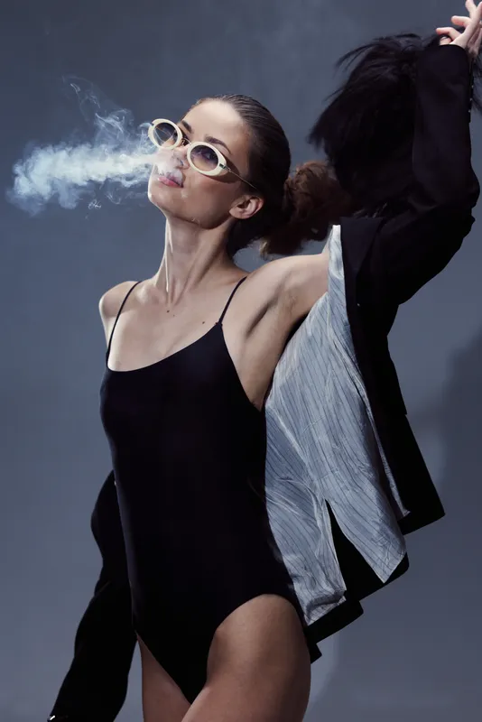 Stylish woman blowing smoke in black bodysuit with sunglasses and loose jacket, moody background and cool tones