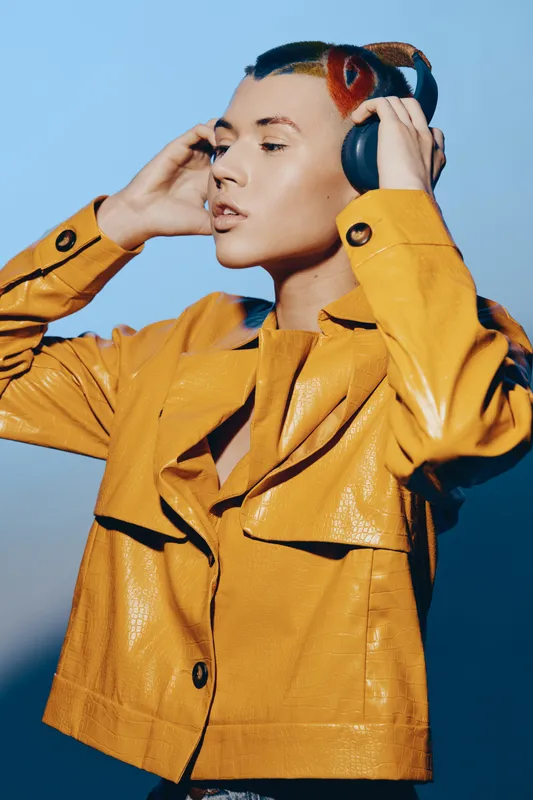young woman wearing yellow jacket listens to music with headphones, stylish look, modern fashion, calm expression, blue background lighting