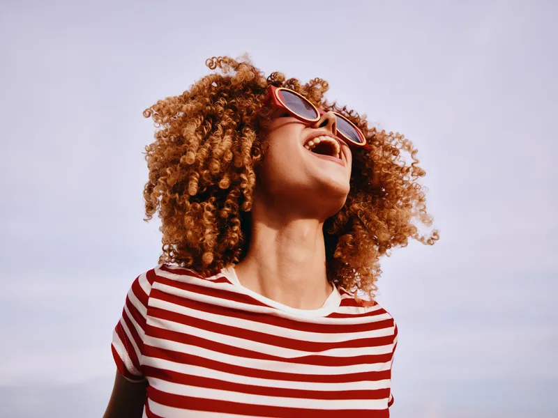A joyful portrait of a woman with curly hair wearing a red and white striped shirt and sunglasses, laughing with an open mouth under a clear blue sky.