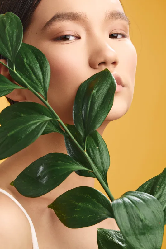 Asian woman portrait with lush green leaves close to her face, presenting serene beauty, natural style, and contemporary elegance in a controlled studio light for a calm, sophisticated mood.