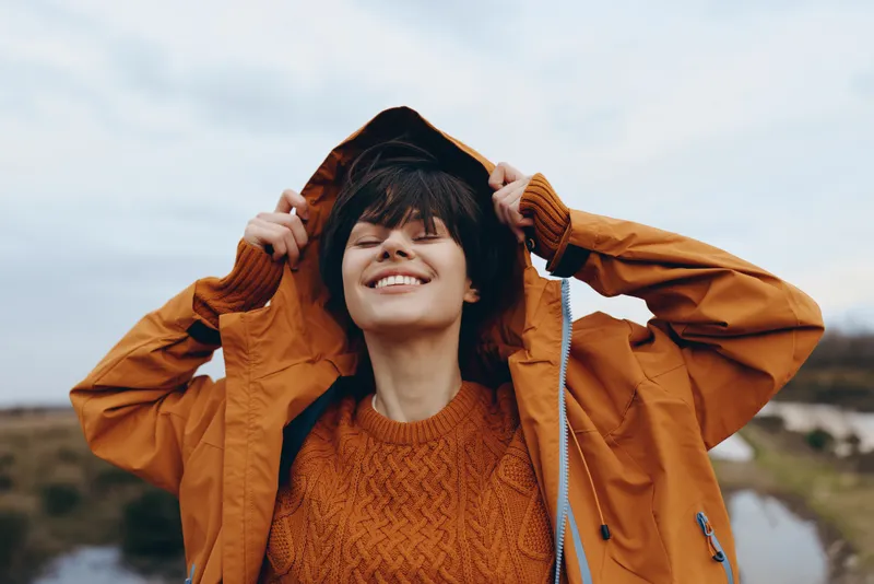 Cheerful woman lifestyle outdoors, smiling as she lifts her jacket hood in cool autumn breeze, soft daylight, river and hills in background, relaxed posture, warm coat, natural colors