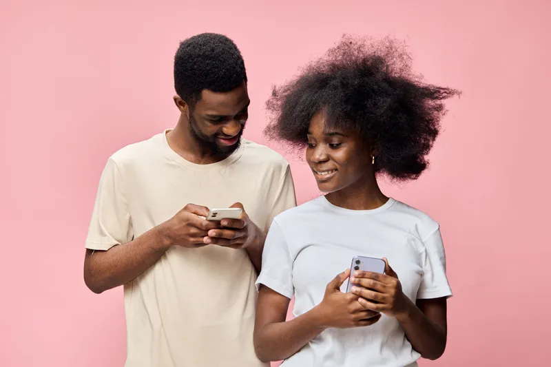 Happy young couple texting on smartphones against a soft pink background, showcasing modern relationships, fashion, and communication