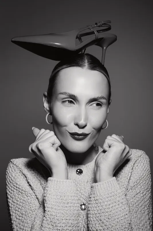 Young white woman with light skin wearing knitted sweater balances high heel shoe on head in black and white studio portrait.