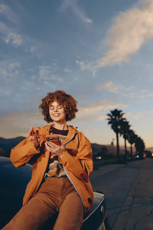 Woman lifestyle moment, smiling with smartphone, curly hair, orange coat, sitting on scooter on city street at sunset, outdoor evening, golden sky, soft clouds, palm trees background