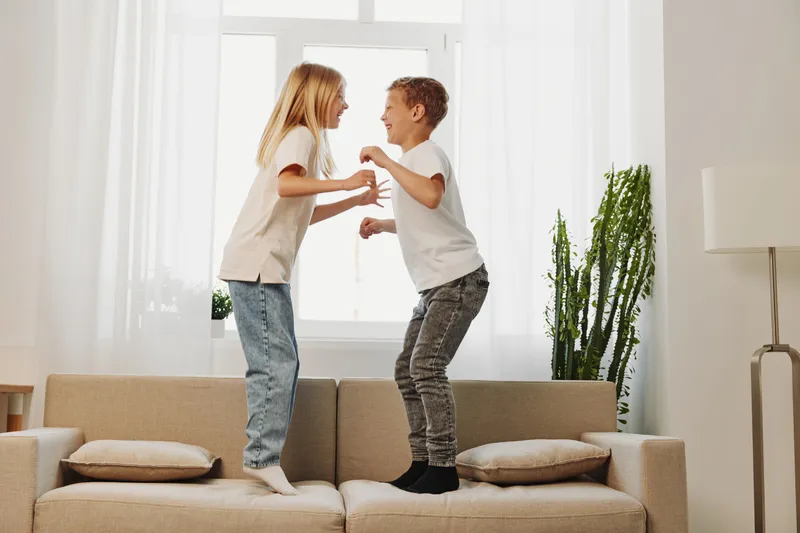 Two children jump on a sofa in a bright living room, smiling and facing each other. The casual home setting includes cushions and a tall green plant by the window.