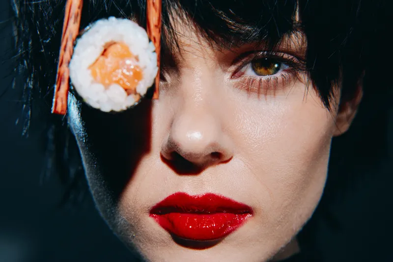 Модная модель с ярким макияжем и суши создает интригующую кулинарную концепцию на темном фоне, подчеркивая современную эстетику в культуре еды