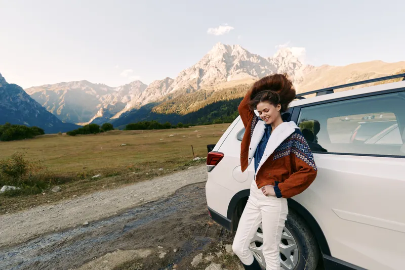Woman leaning on car by mountains and landscape on a gravel road, travel portrait in nature wearing warm jacket, casual SUV trip with sunlight and relaxed holiday vibe