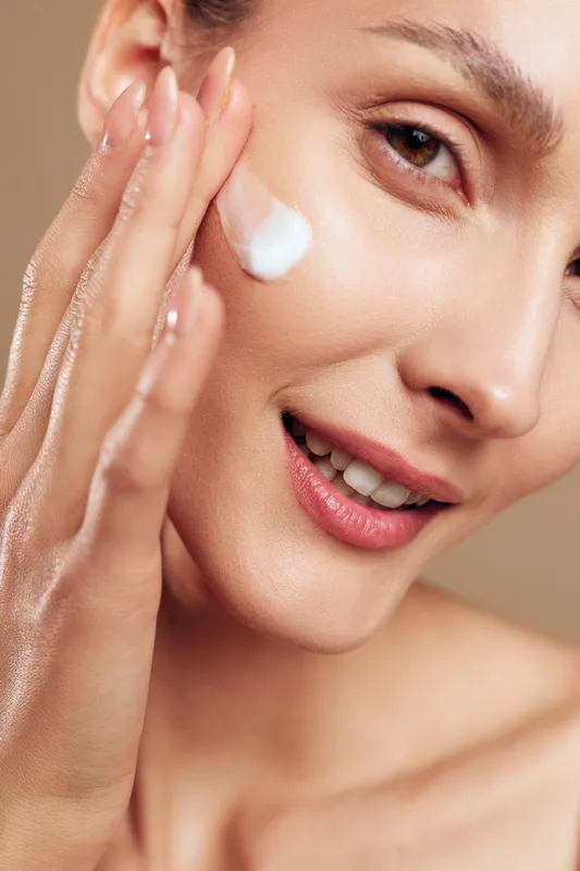 Young woman with clear skin gently applies white face cream on her cheek while smiling softly against a beige background, conveying a sense of skincare and self-care.