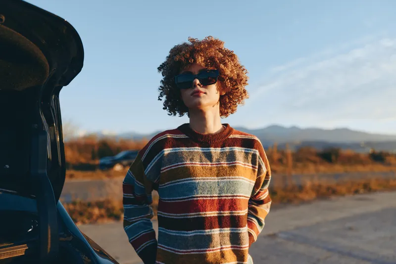 Woman lifestyle curly hair sunglasses sweater, standing by car at sunset outdoors, warm light golden landscape autumn hills, side view scenic countryside road, relaxed casual portrait