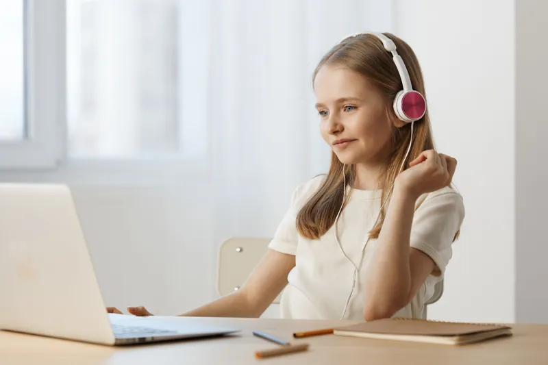 Young girl with headphones enjoying music while using a laptop in a bright, minimalist workspace, reflecting happiness and focus in a modern setting