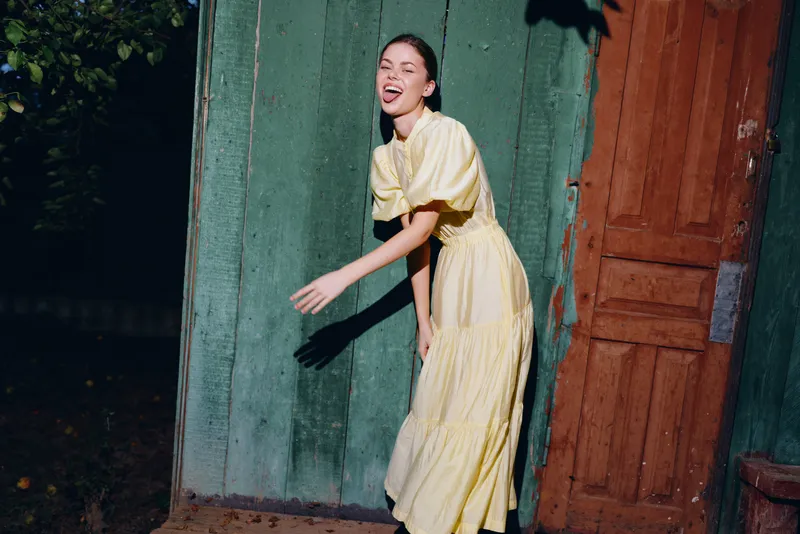 woman in a yellow dress playfully leans against a colorful wall outdoors capturing a candid moment with bright light and a carefree expression showcasing vibrant street style