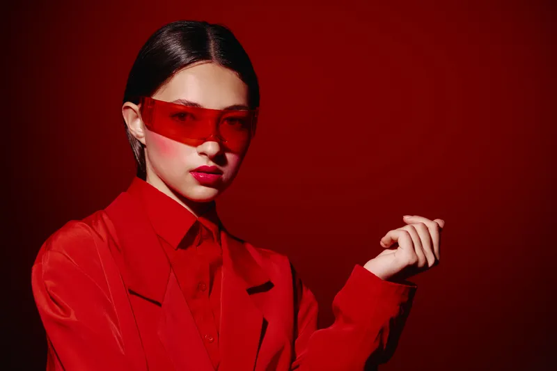 Stylish young woman wearing oversized red translucent sunglasses and a shiny red jacket poses against a deep red background. Bold color contrast emphasizes fashion and beauty.