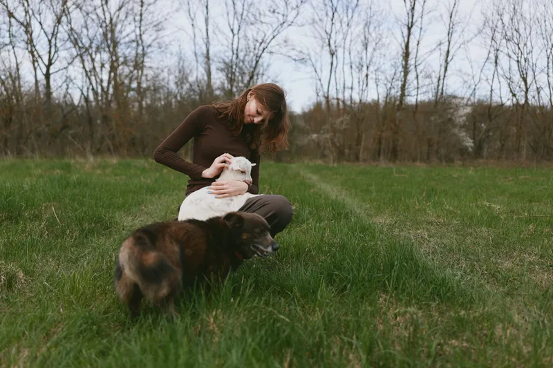 Young woman with long hair sits on grass in a field gently holding a white baby goat while a brown dog stands nearby in a calm outdoor lifestyle scene.