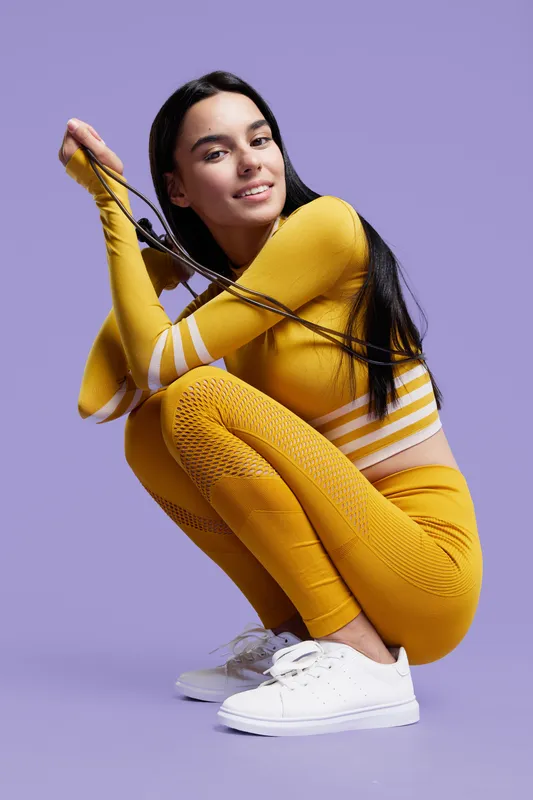 Sport fitness woman in yellow activewear squatting and smiling, holding jump rope against purple background, young athletic female demonstrating workout readiness.