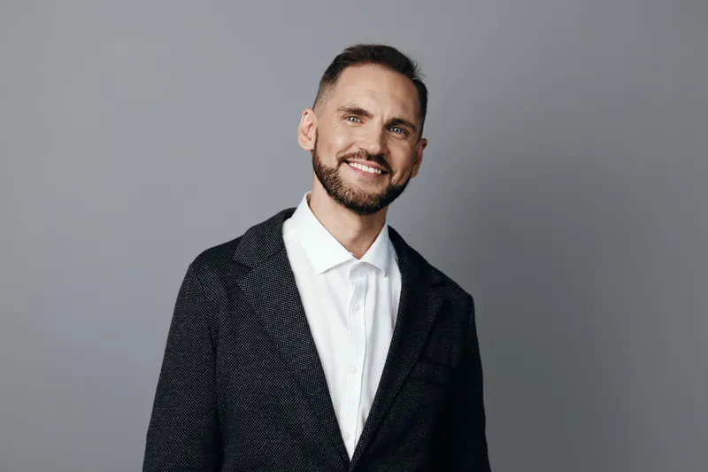 A professional man in a sharp blazer on a solid gray backdrop, radiating confidence and friendly charm. This corporate portrait conveys poised leadership and approachable demeanor.