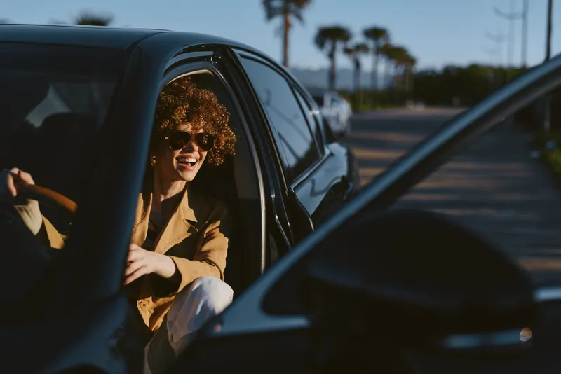 Smiling woman with curly hair in a car, daylight streaming through the window. Retro film color, casual road trip mood, urban street background, warm and relaxed lifestyle.