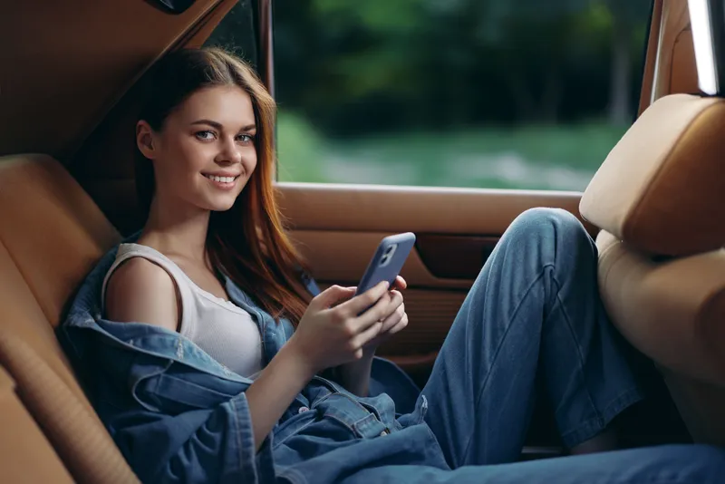 Young woman smiling while using a smartphone in a car, wearing casual denim attire, expressing joy and relaxation in a natural green background.
