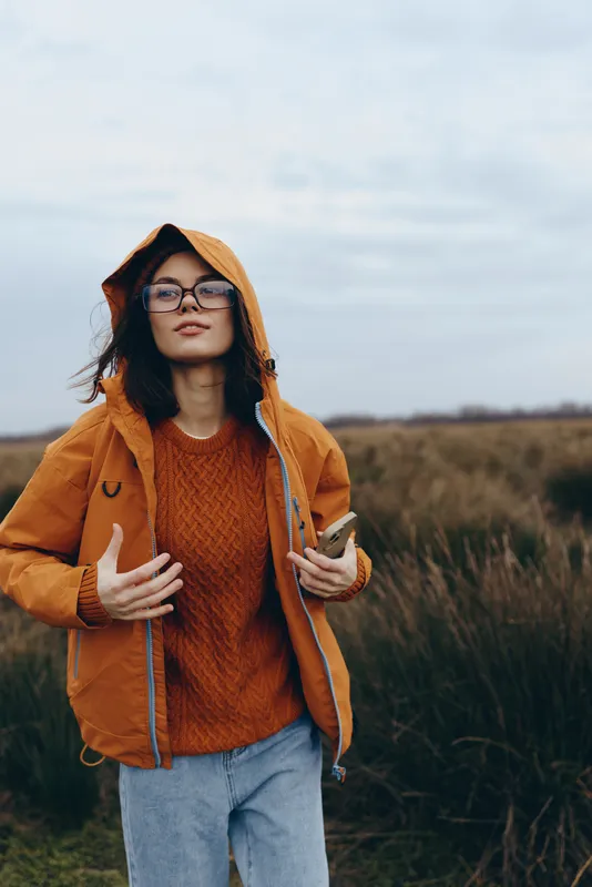 Woman outdoors in autumn field, orange hoodie and knitted sweater, glasses, hood up, relaxed lifestyle pose, gentle wind, soft diffused natural light, warm colors, tall grass background