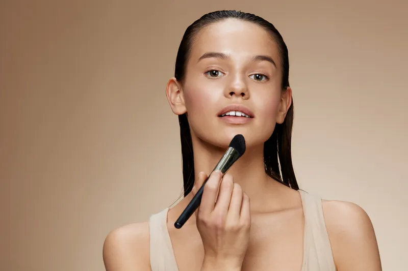 Young model applying makeup with a brush, showcasing a natural look and confident expression against a warm beige background, reflecting beauty and self care