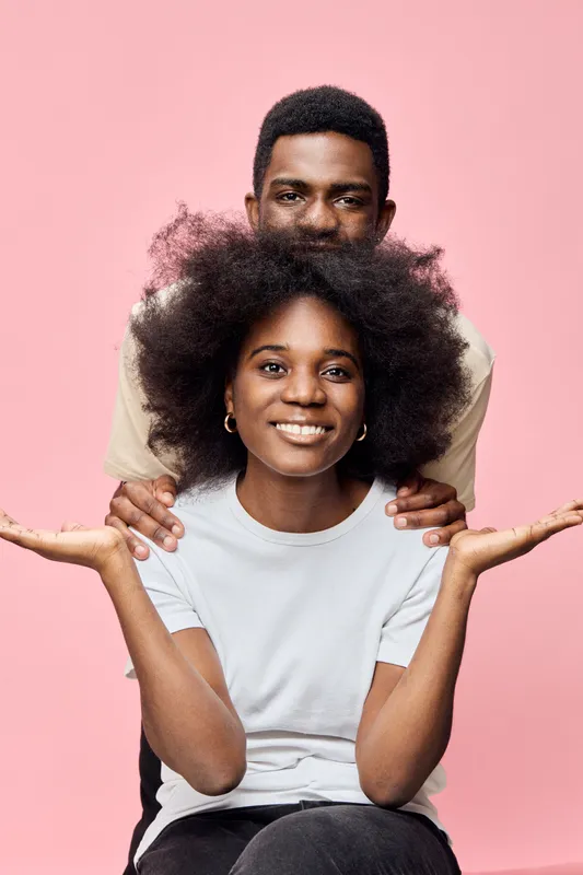 A joyful young African couple posing together, showcasing cheerful expressions, vibrant costumes, and a soft pink background with a light hearted aura