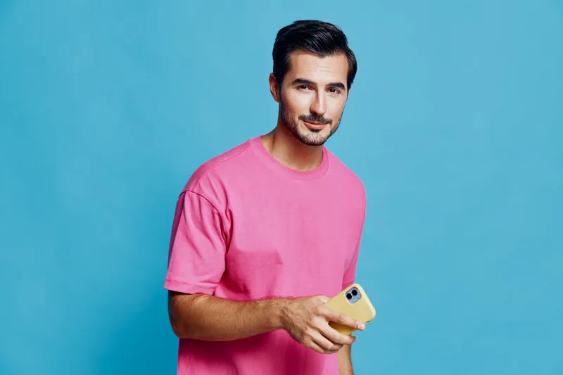 Full body smiling man wearing a pink t-shirt looks at the camera while holding a yellow smartphone against a vibrant blue background. Modern lifestyle concept.