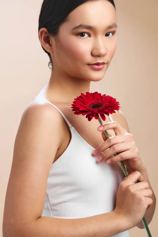 asian woman in a white sleeveless top stands in a studio with soft light, smiling gently while holding a red flower near her chest, exuding calm grace and natural beauty.