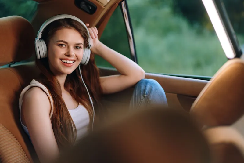 Young woman enjoying music while sitting in a car, wearing headphones and smiling, surrounded by a green outdoor background, conveying a carefree and joyful mood.