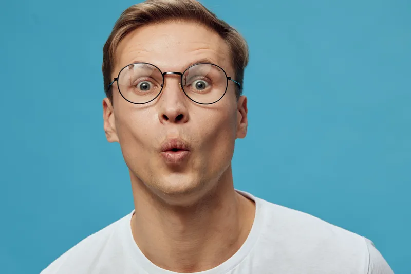 Close-up of surprised young Caucasian man with glasses, unexpected expression, wearing white T-shirt, isolated on bright blue background, studio portrait, people lifestyle concept, humorous mood, faci