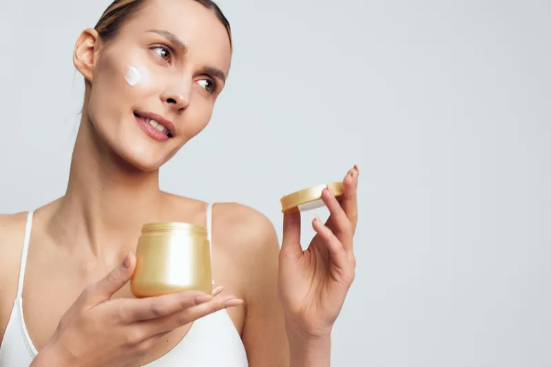 Young woman with clear skin holding open jar of cream applying moisturizer on face, natural beauty concept, soft lighting, neutral background, skincare routine moment