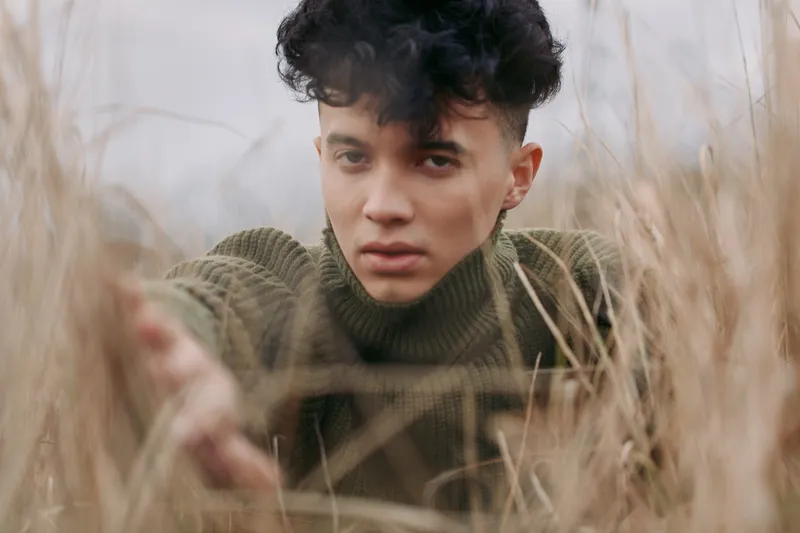 A close, intimate portrait of a young person reaching through tall dry grass, raw expression and natural light conveying a sense of authenticity and credibility in a quiet outdoor moment.
