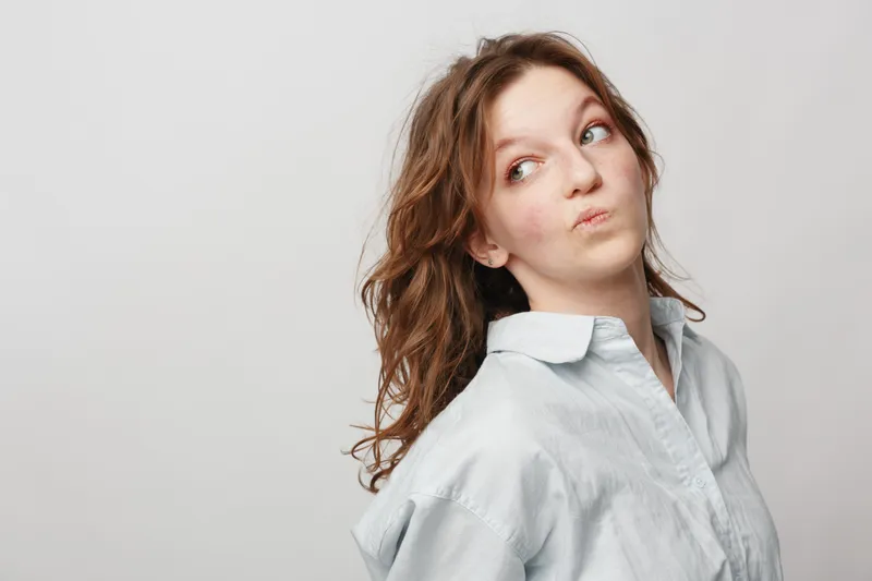 Young woman with curly hair wearing a light shirt looks sideways with a thoughtful expression on a plain white background. Concept of contemplation, curiosity, and casual portrait.