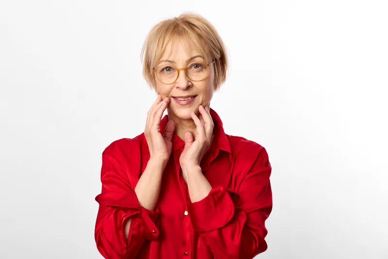 Elderly woman with glasses in a red blouse studio portrait, hands at her face show surprise and a warm smile on a plain white background, concept for positive emotion and happiness
