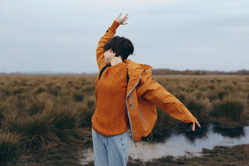 Woman outdoors in orange jacket and jeans, seen from behind in windy field. Walking with raised hand, lifestyle and nature fit this free mood under cloudy sky.