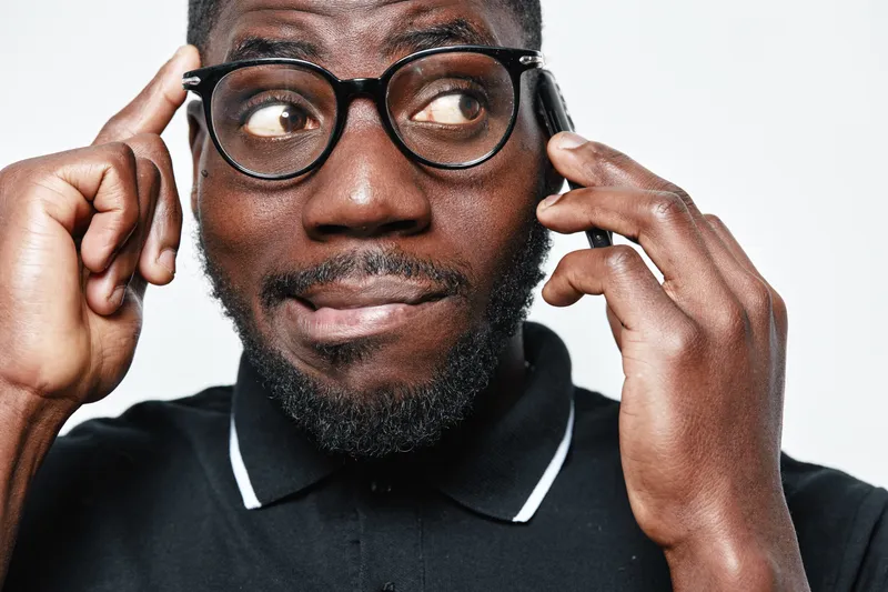 African American man with dark skin and glasses talking on smartphone, thinking and smiling in casual black polo shirt on white background. Closeup portrait of confident male.