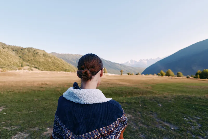 Woman back in sweater stands in open field overlooking mountain landscape and distant valley, travel scene with solitude and nature view, outdoor exploration and calm horizon.