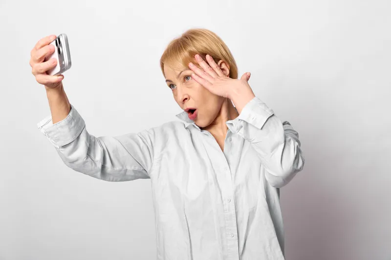 Woman holding smartphone taking selfie, surprised elderly expression, hand covering forehead, face reaction, studio portrait on light gray background, soft diffused lighting, indoor