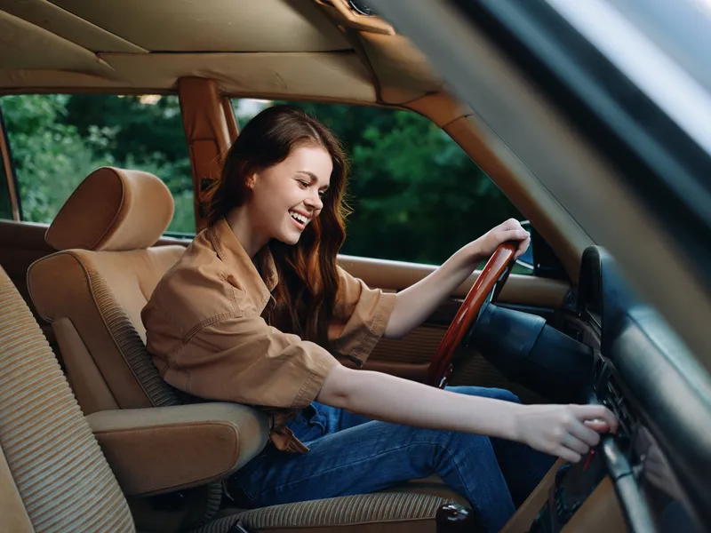 Happy young woman driving a vintage car, enjoying freedom and adventure, dressed in casual attire with a warm color palette and a natural background.