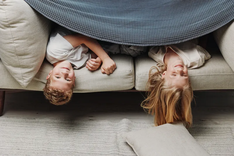 Two happy children with different hair colors lie upside down on a sofa under a grey blanket, smiling and playing in a cozy living room environment.