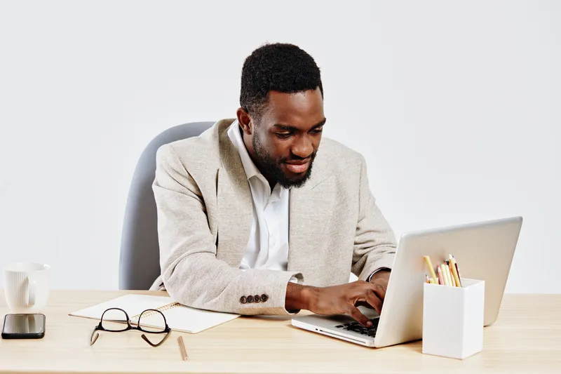Professional African American man working on laptop at desk in modern office, wearing beige blazer and white shirt, focused on screen, workspace organized with glasses, mug, and stationery, business a