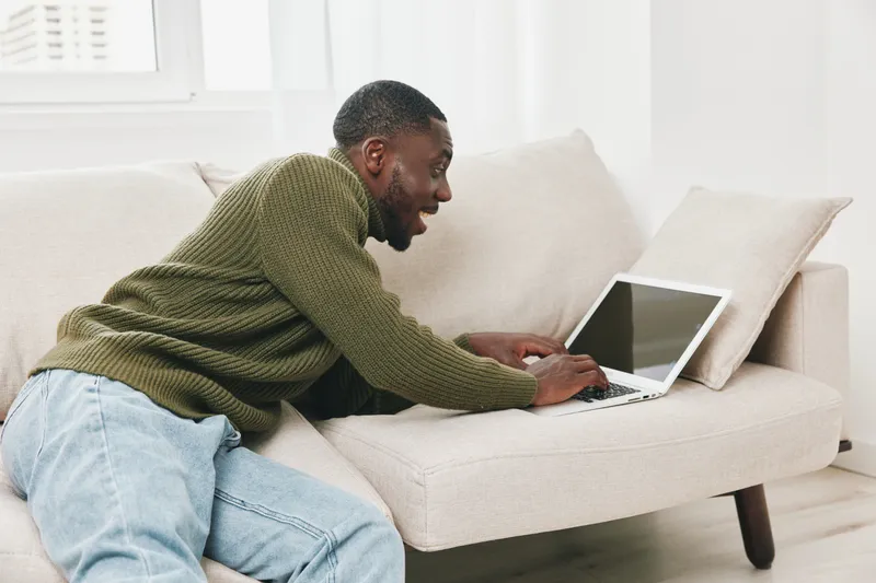 Young Black man excitedly using a laptop on a cozy couch, wearing a green sweater and jeans in a bright room, showcasing emotions of joy and engagement