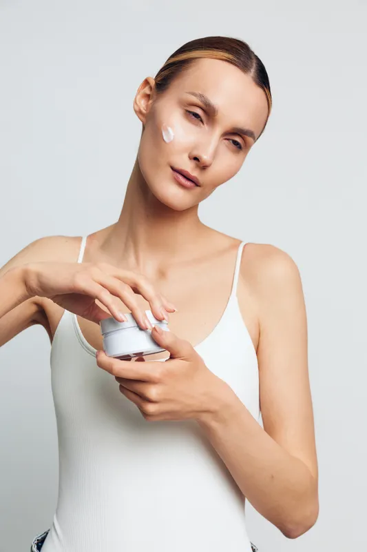 A young woman in a white tank top applies cream to her face with her eyes closed. The neutral background emphasizes skincare, beauty routine, and self care concepts.