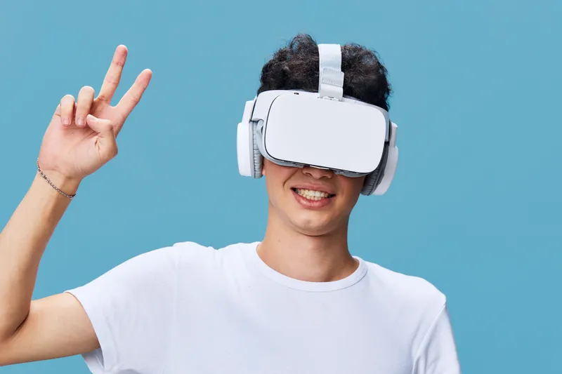 young man wearing virtual reality headset smiling and showing peace sign with hand against blue background, casual clothing, technology experience