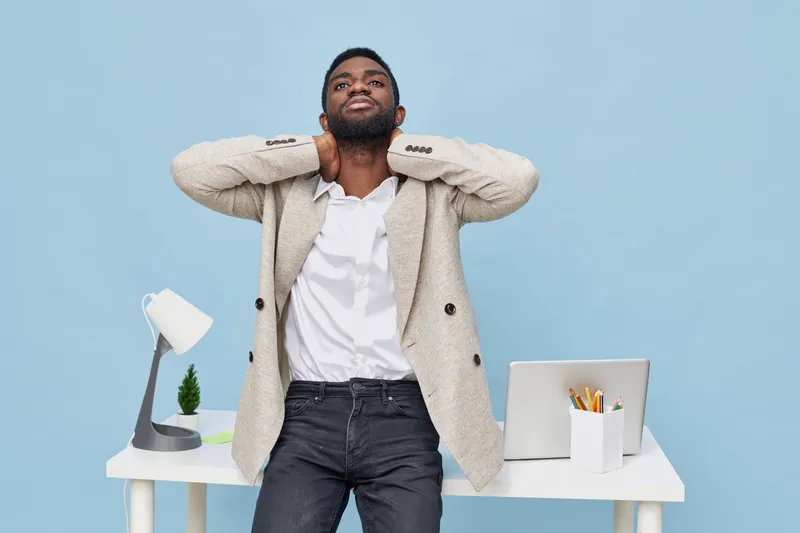 Relaxed African American man in casual outfit stretchs neck while standing behind modern white desk with lamp and laptop against blue background, studio shot, lifestyle concept, relaxation and self-ca