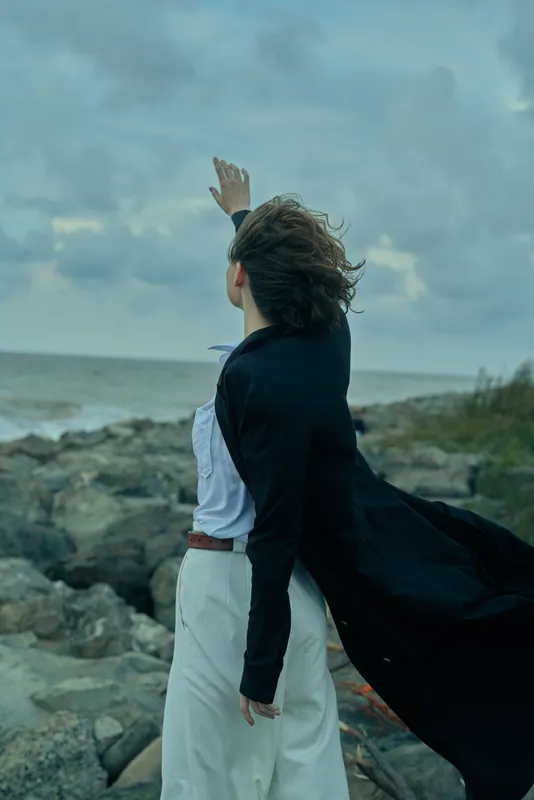 Woman on a rocky shoreline raises her arm toward the sky, cape billowing in the wind, capturing a moment of freedom, determination and dramatic outdoor scene.