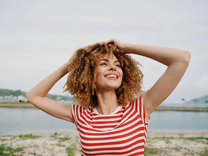 Joyful portrait of a woman with curly hair wearing a red and white striped shirt, outdoors by a calm lake, catching sunlight and radiating carefree confidence.